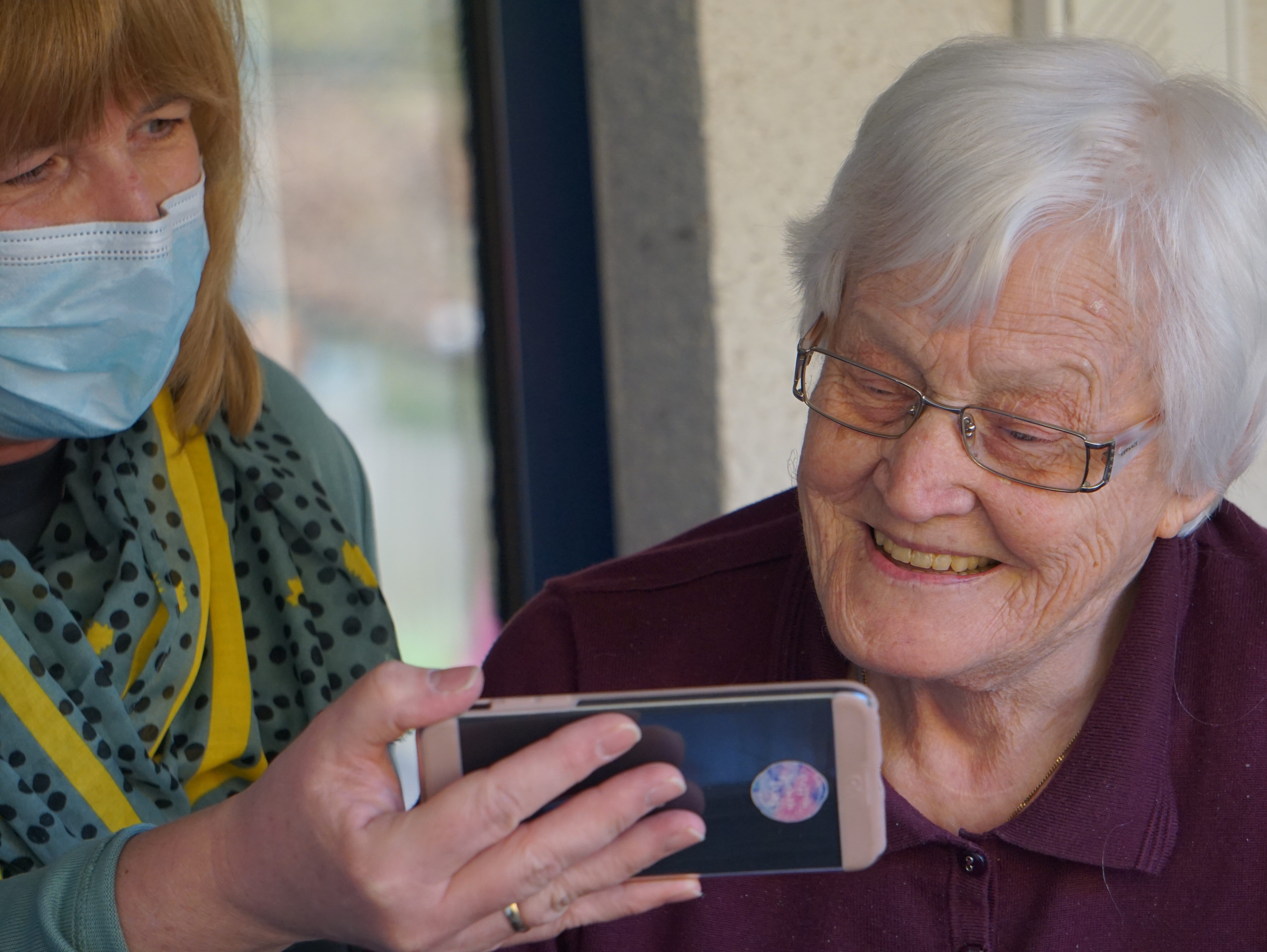 A woman in a mask showing a smiling older lady something on a mobile phone.