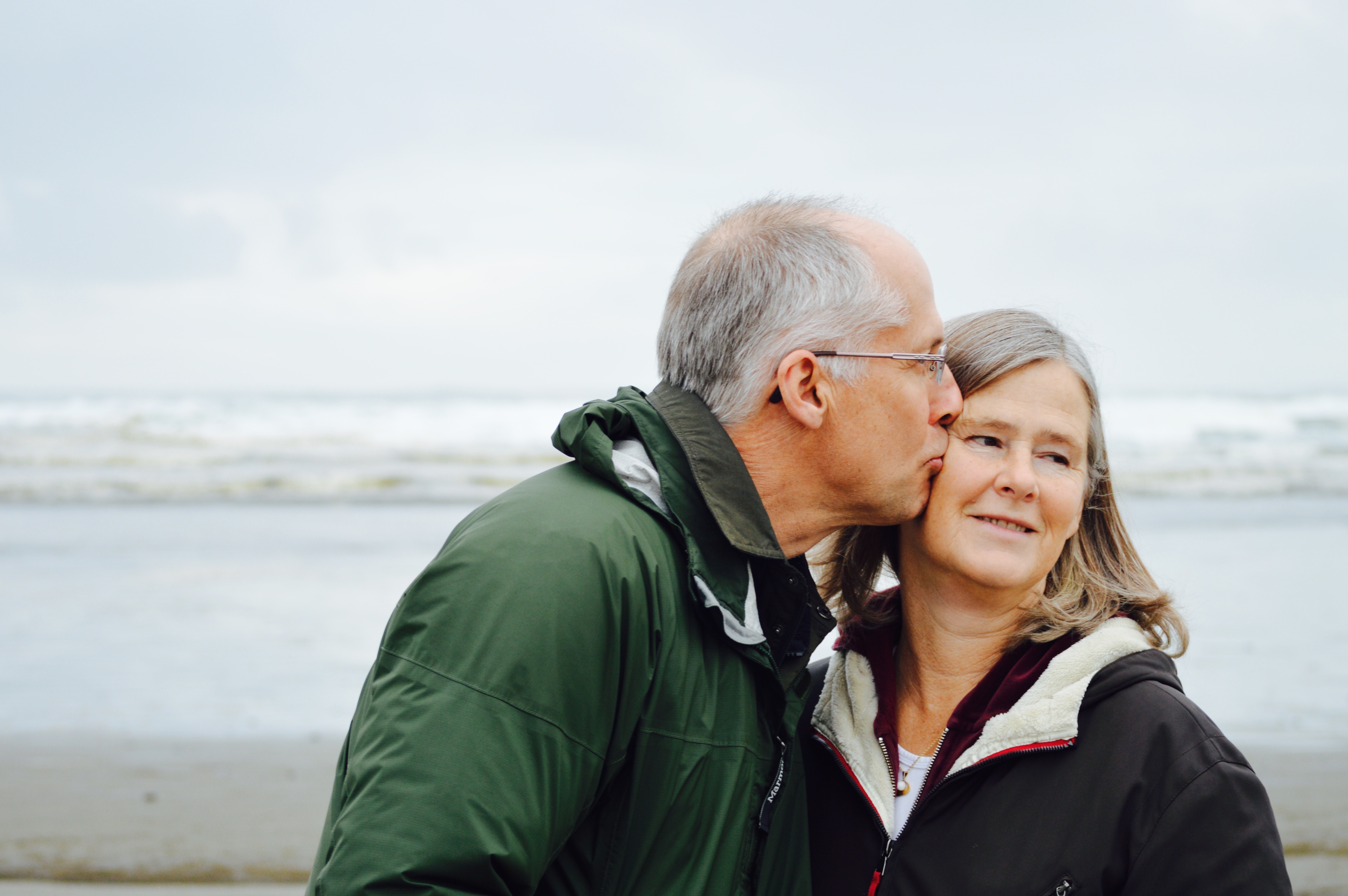 An older couple - a man and a woman - kissing on a beach wearing anoraks.