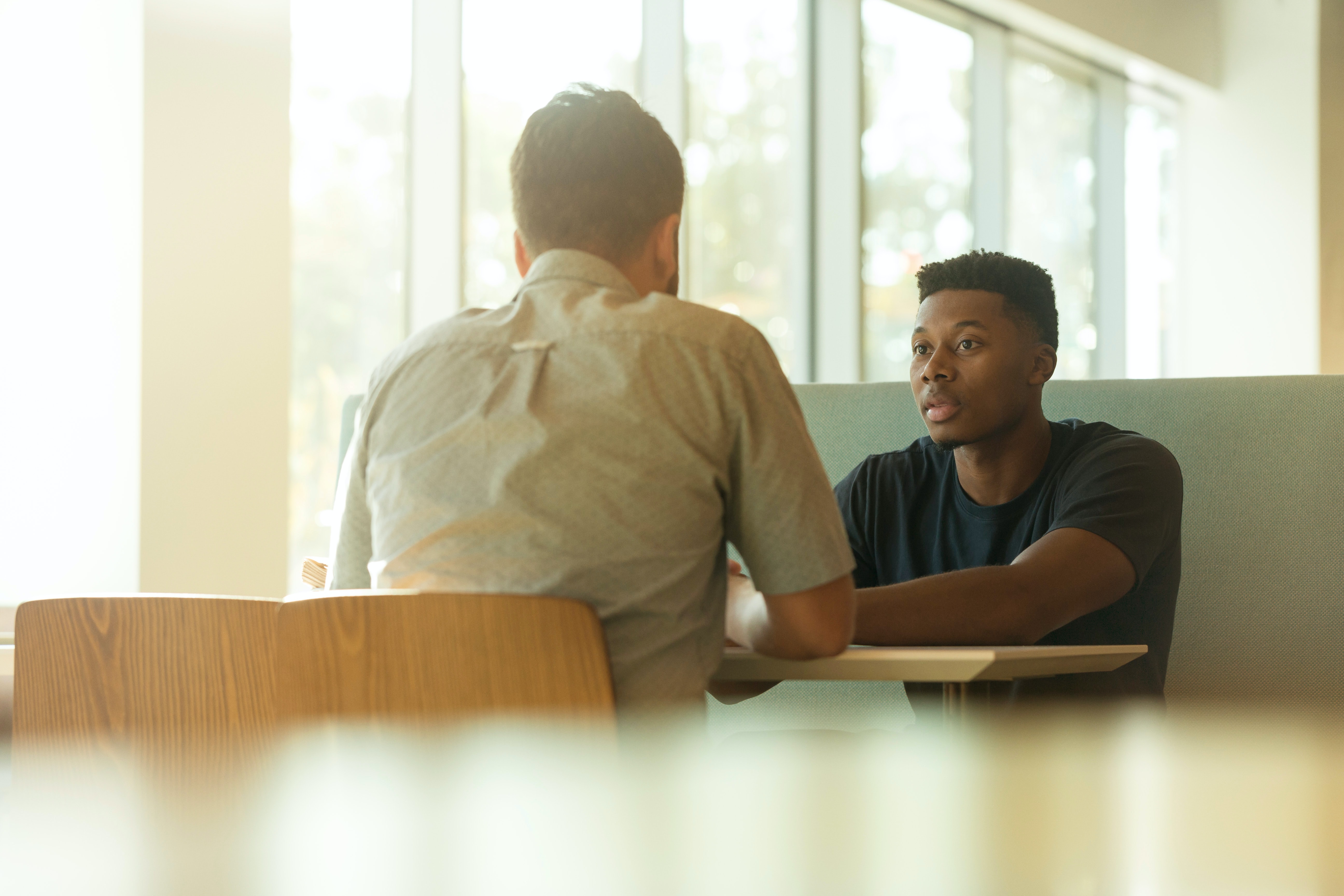 A young man talking to an older man across a table in a sunny office.