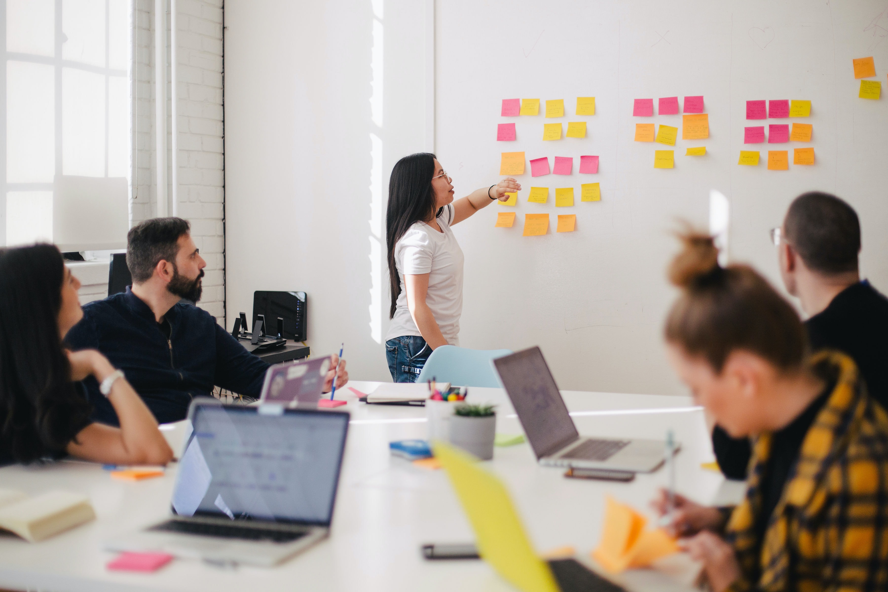 A group of people working together around a large white table while a young woman adds a post it note to a wall of coloured post it notes.