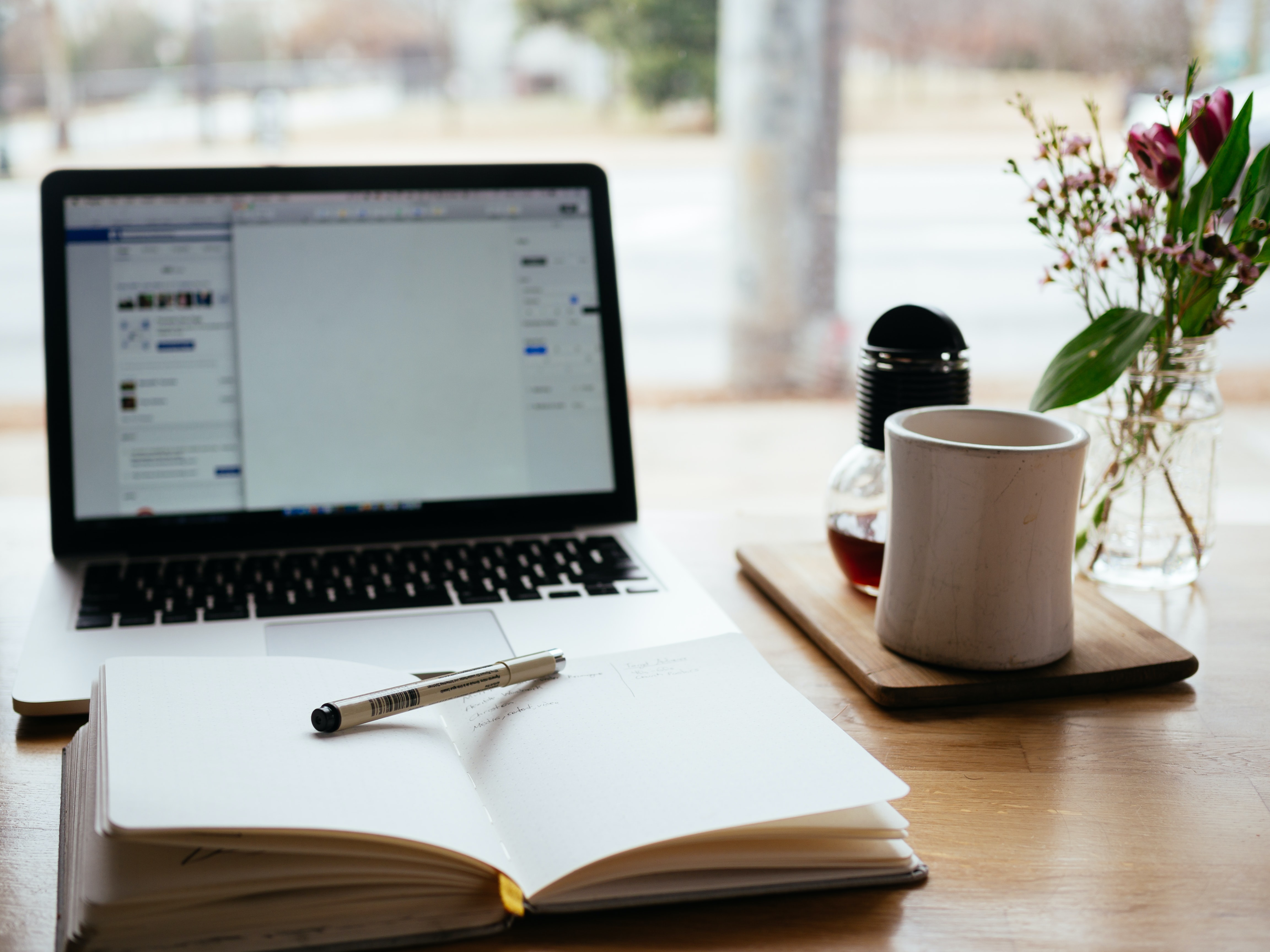 A notebook and a laptop on a desk with a pen and a cup of coffee