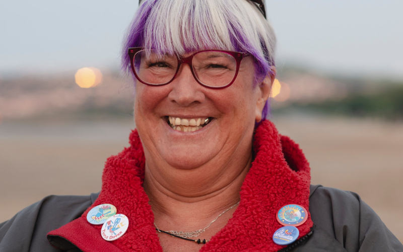 An older lady smiling positively at the camera, with a jacket on. There are 4 badges on her jacket and she is stood on a beach.